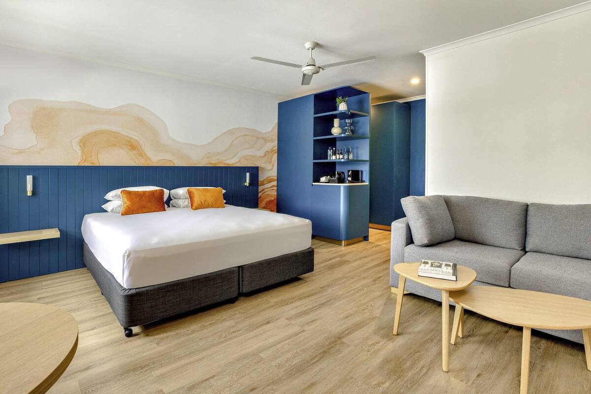A modern hotel room with a bed, a sofa, a small table, and a built-in shelf unit.