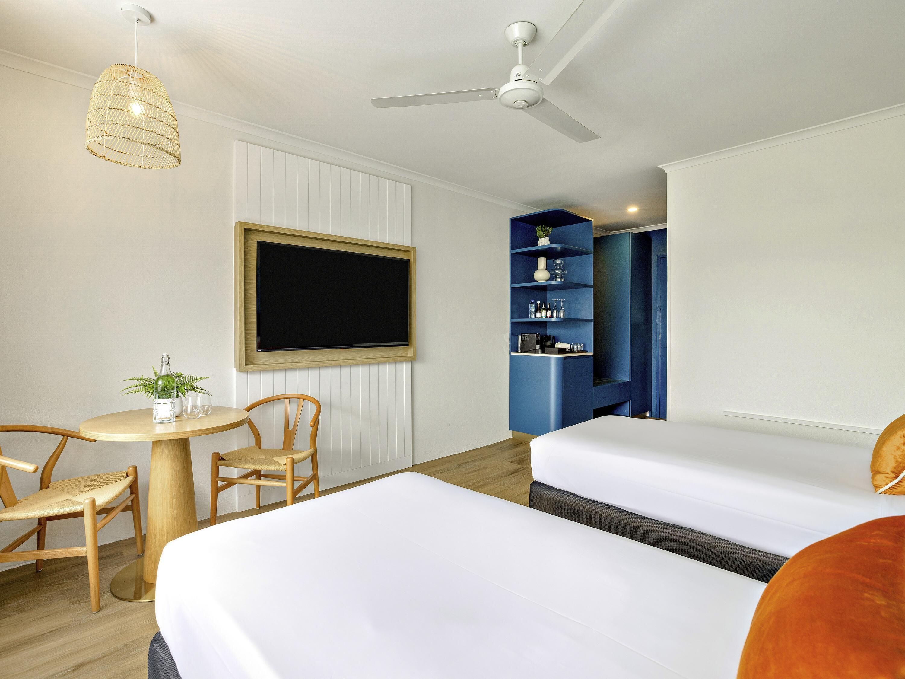 Habitación de hotel con cama, escritorio, silla, ventilador de techo, televisor montado en la pared y estantería azul.