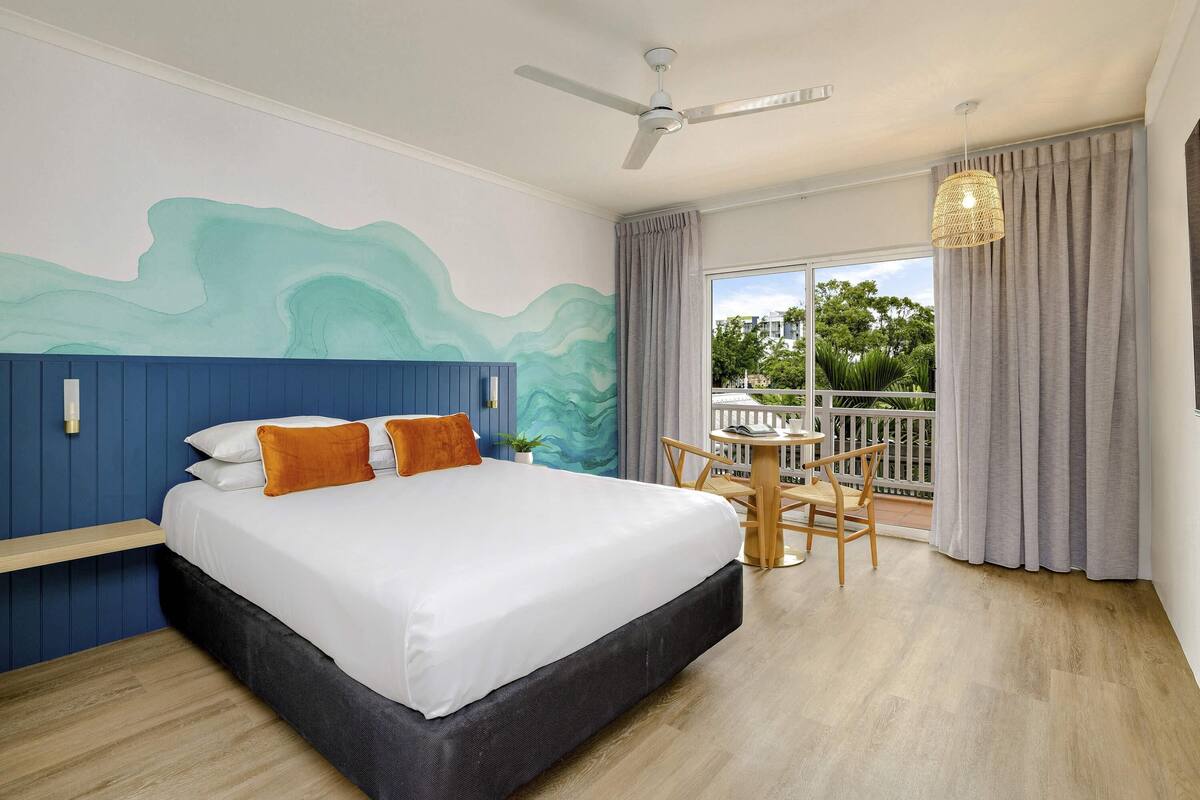 A bedroom with a bed, a ceiling fan, a balcony with a table and chairs, and a view of greenery outside.