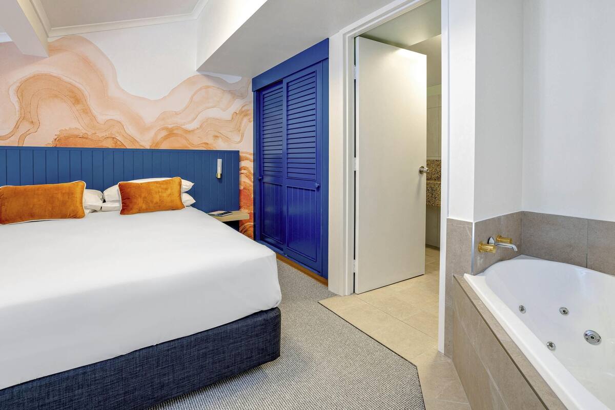 A modern hotel room with a large bed, a blue door, a bathroom with a bathtub, and a marble wall.