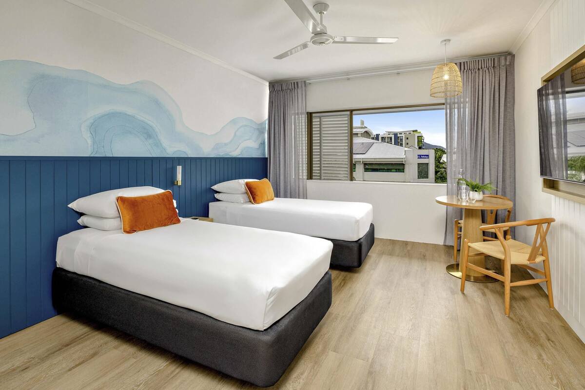 A hotel room with two beds, a ceiling fan, a small table with chairs, and a view of buildings through the window.