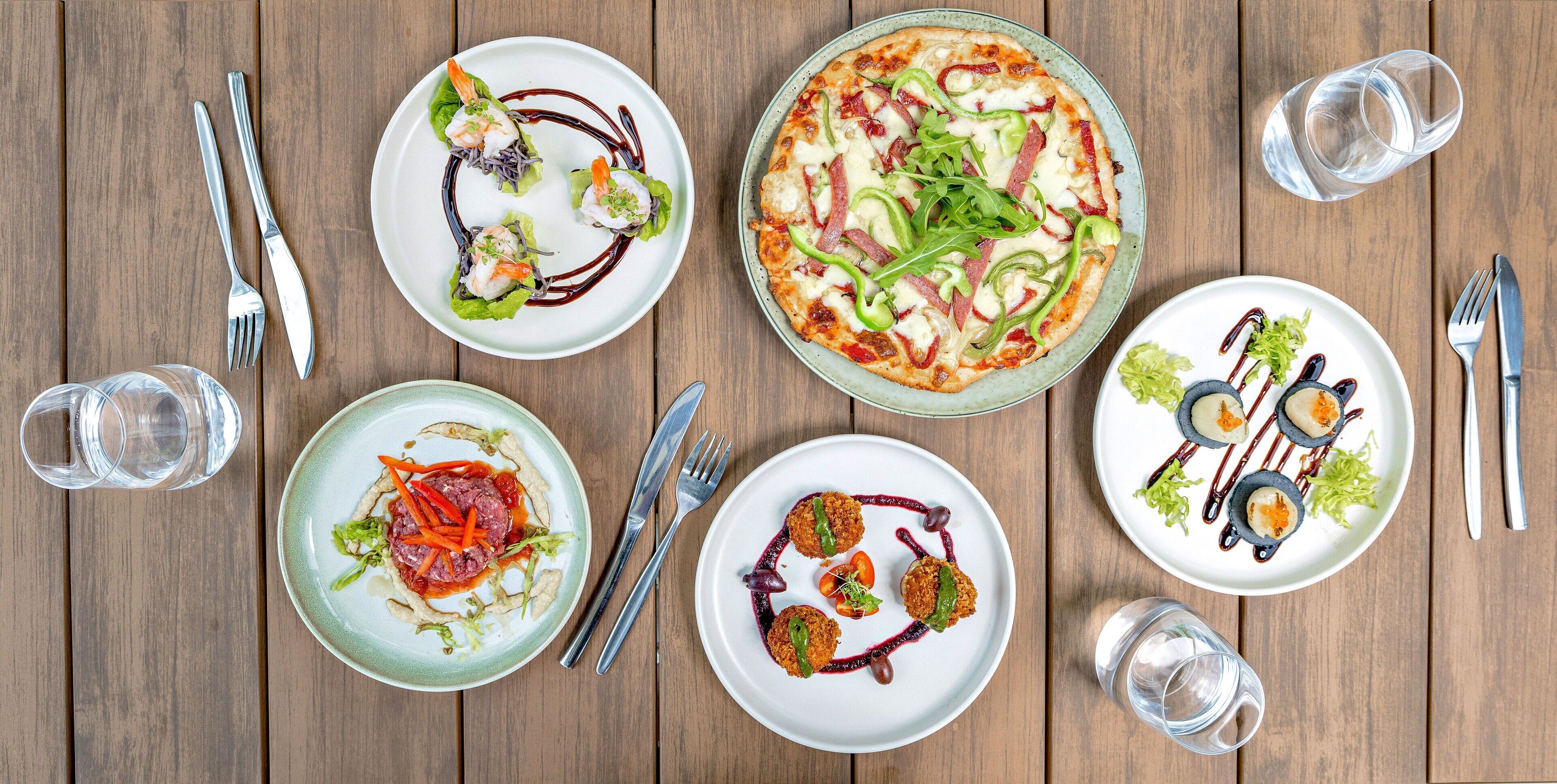 Ein Holztisch mit verschiedenen Gerichten, darunter Pizza, Salate und Vorspeisen, für das Essen angerichtet.