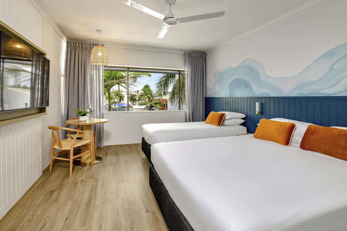 A hotel room with two beds, a ceiling fan, a TV, a small table with a chair, and a window with a view of palm trees.