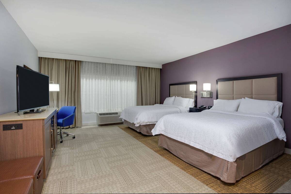 A hotel room with two beds, a desk with a flat-screen TV, a chair, and a desk lamp.