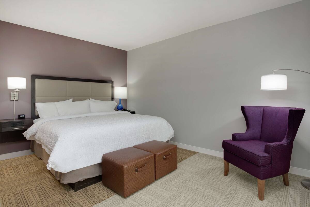 A hotel room with a bed, two bedside tables, a purple armchair, a floor lamp, and a small chest of drawers.