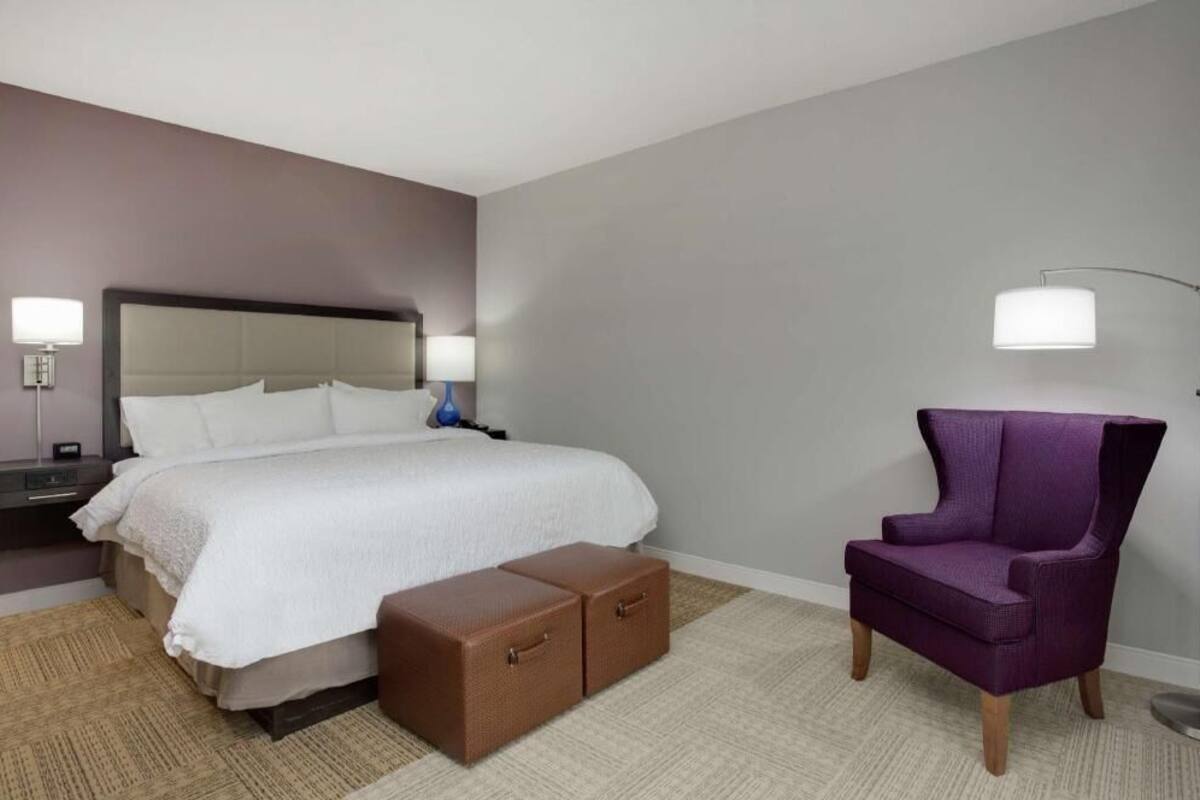 A hotel room with a bed, bedside tables, a lamp, a purple armchair, and a small chest of drawers.