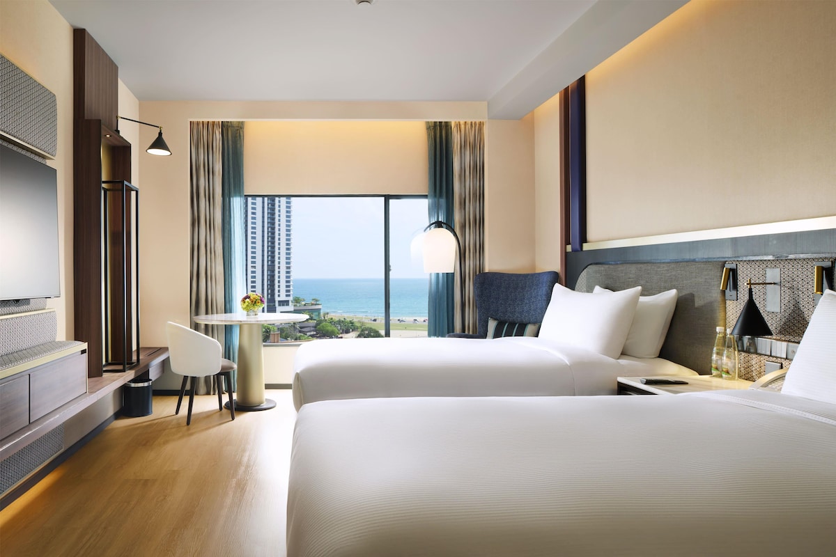 Habitación de hotel con dos camas, un escritorio, una silla y vista al océano.