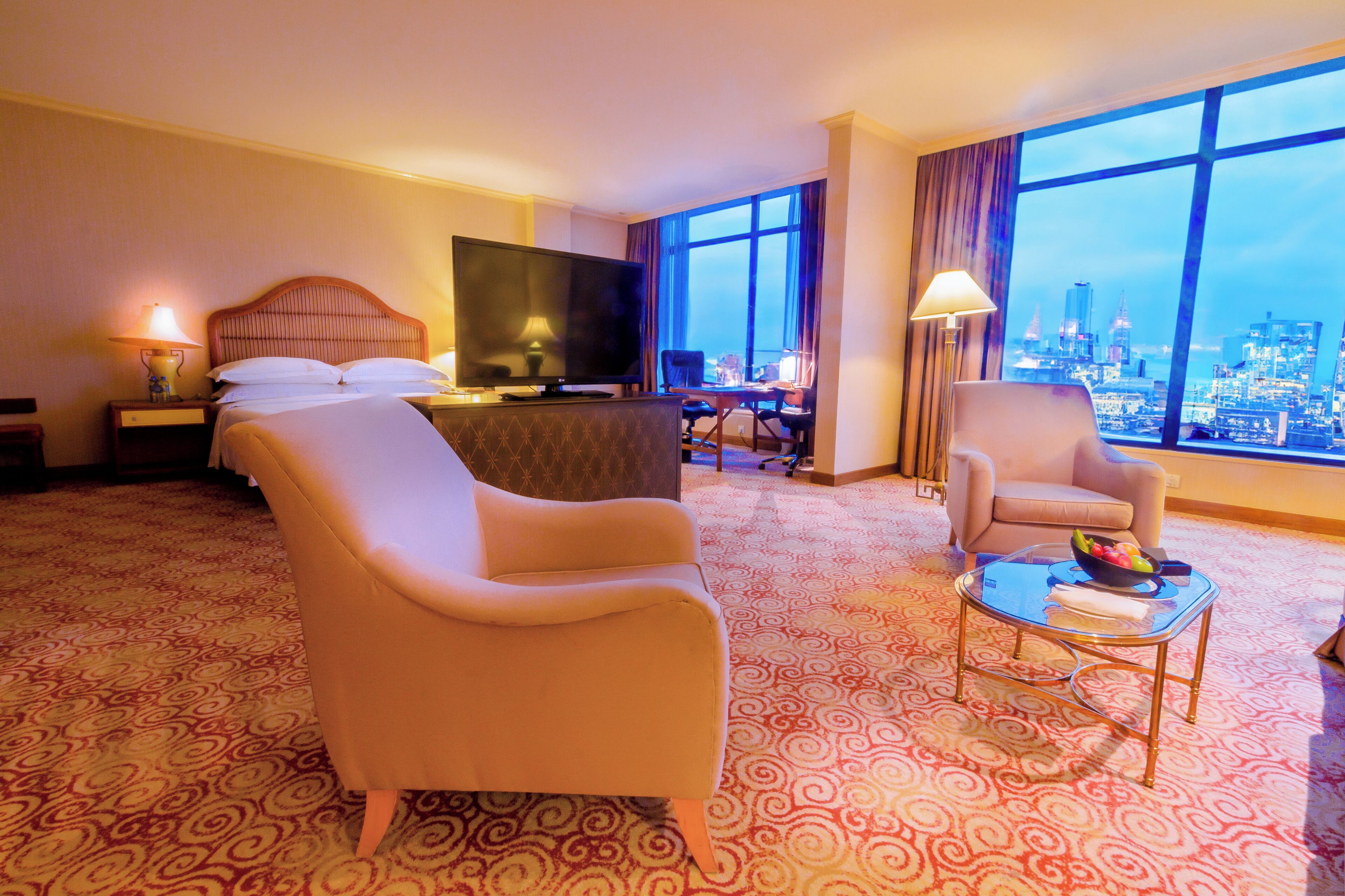 Habitación de hotel con cama, televisión, escritorio, dos sillones y vista a un paisaje urbano a través de ventanas amplias.