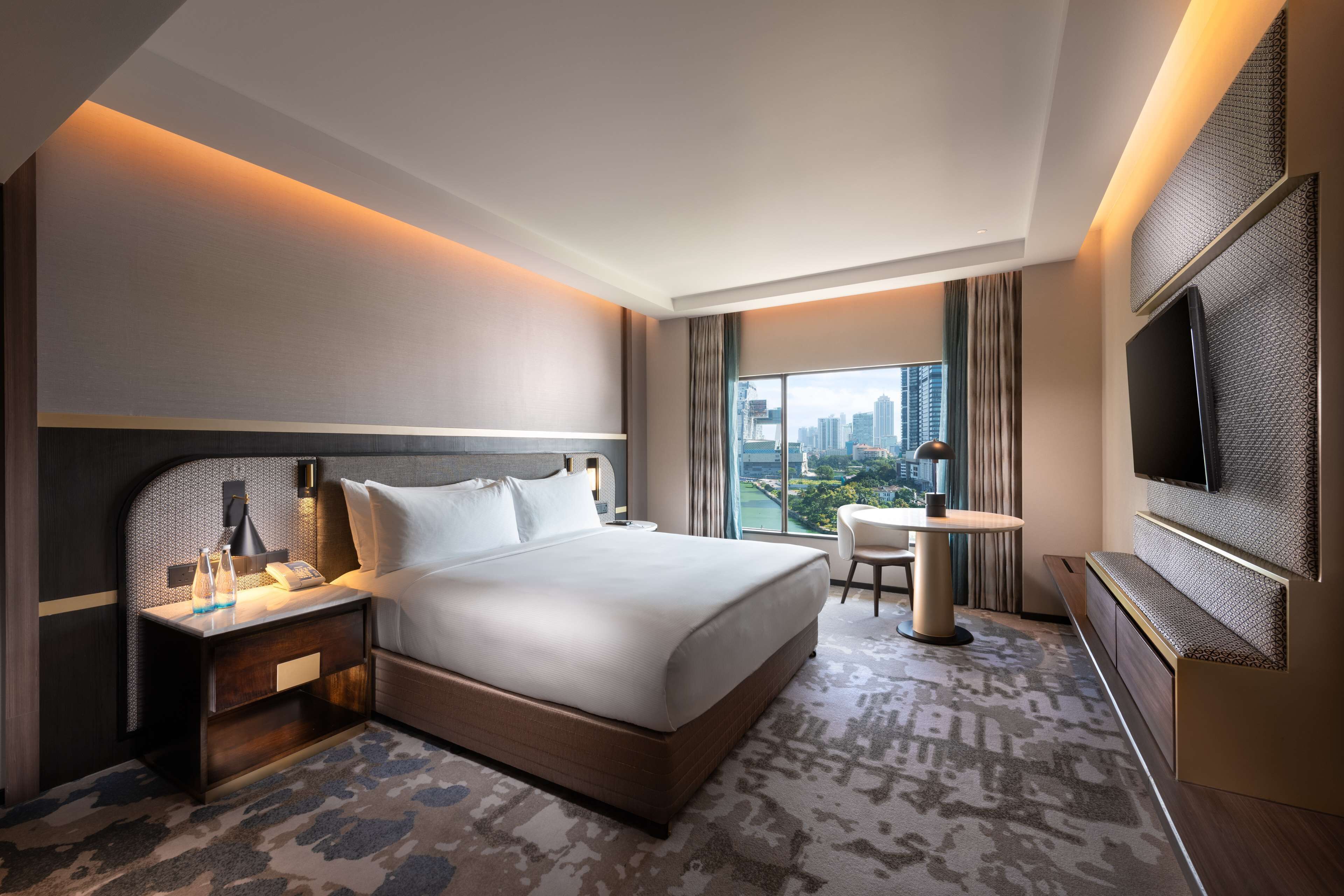 A modern hotel room with a large bed, a desk, a chair, a TV, and a city view.