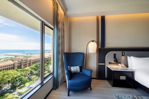 Habitación de hotel con un ventanal grande, un sillón azul, una mesita de noche con lámpara y una cama.