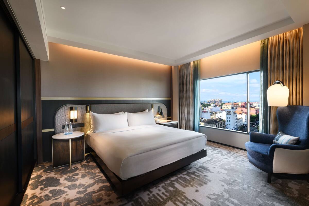 Ett modernt hotellrum med en stor säng, en sittgrupp med en fåtölj, ett nattduksbord och utsikt över staden.