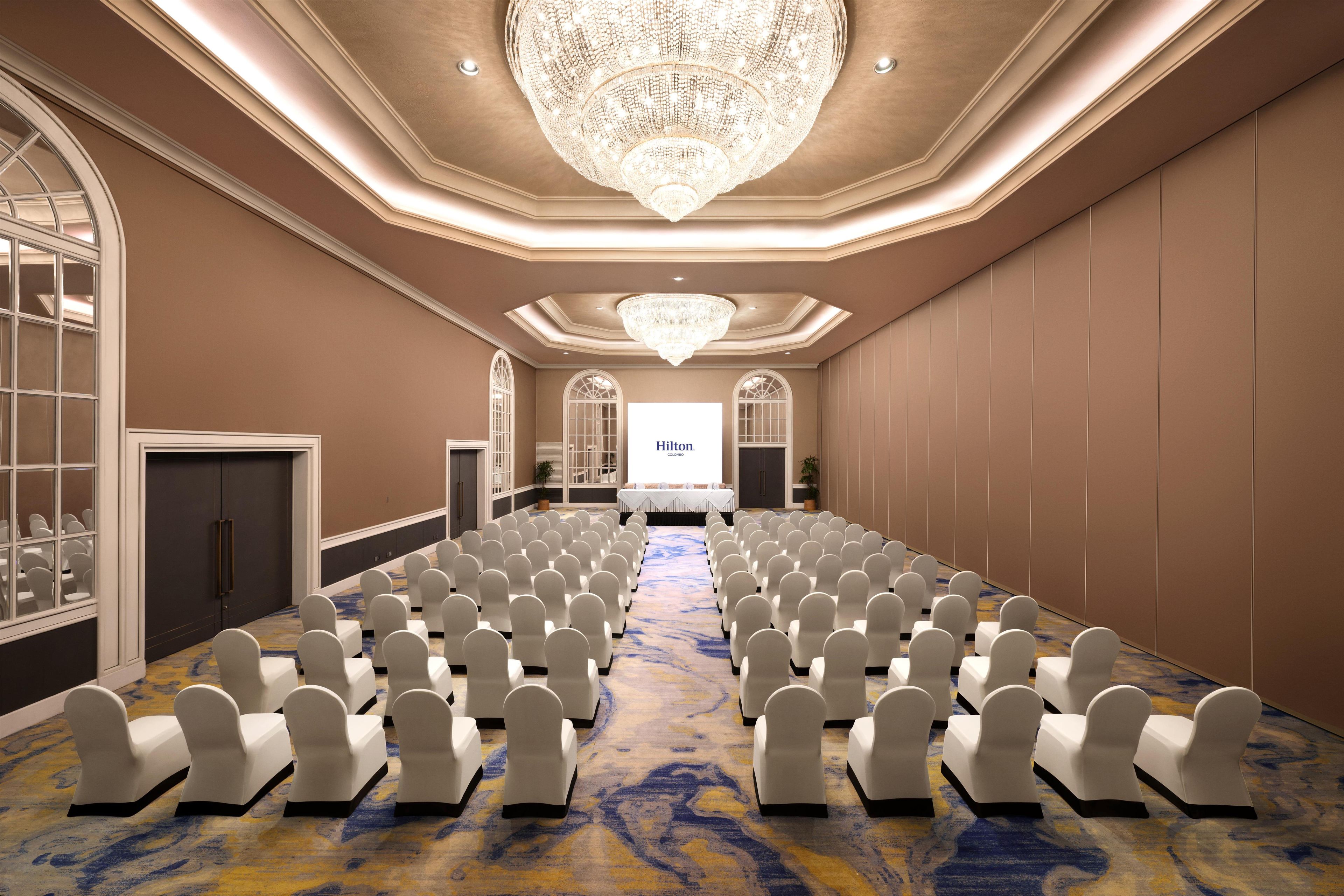 Une salle de conférence avec des rangées de chaises, un grand lustre et un écran de présentation.
