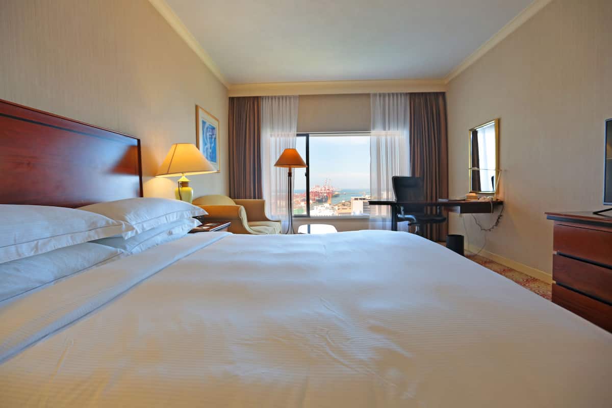 A hotel room with a large bed, a desk, a chair, a lamp, and a view of a bridge.