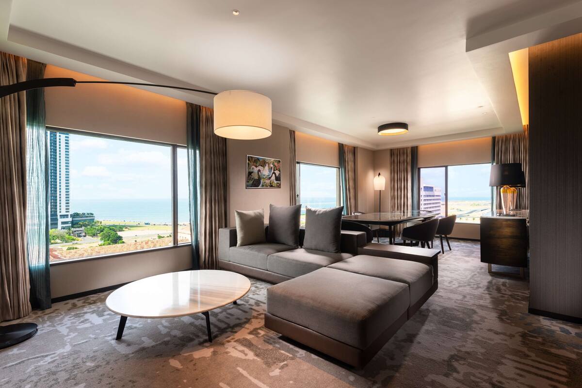 Ett modernt hotellrum med ett stort fönster som erbjuder utsikt över stranden och havet, en soffa, ett matbord och ett litet soffbord.