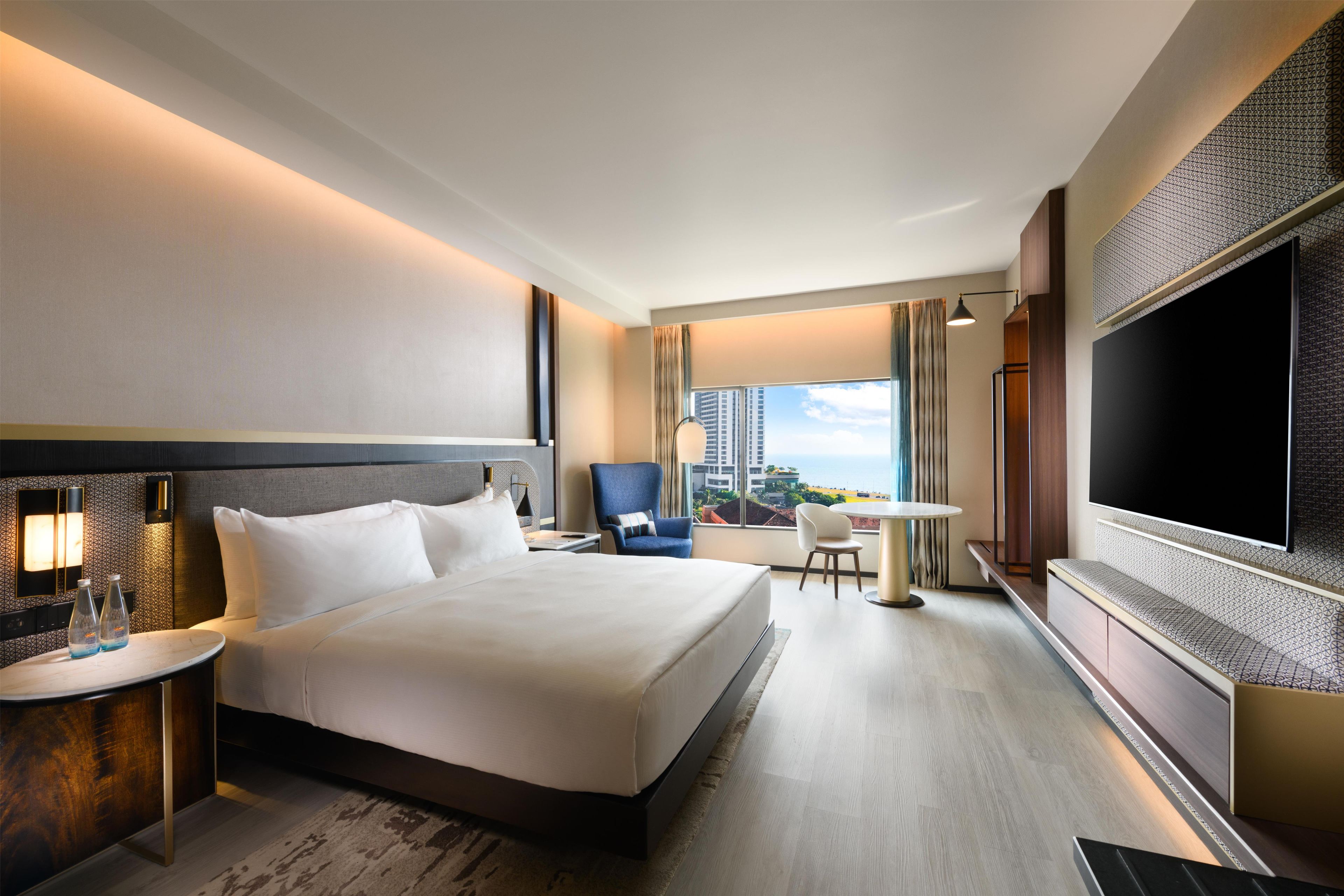 A hotel room with a large bed, a desk, a chair, a TV, and a view of the city.