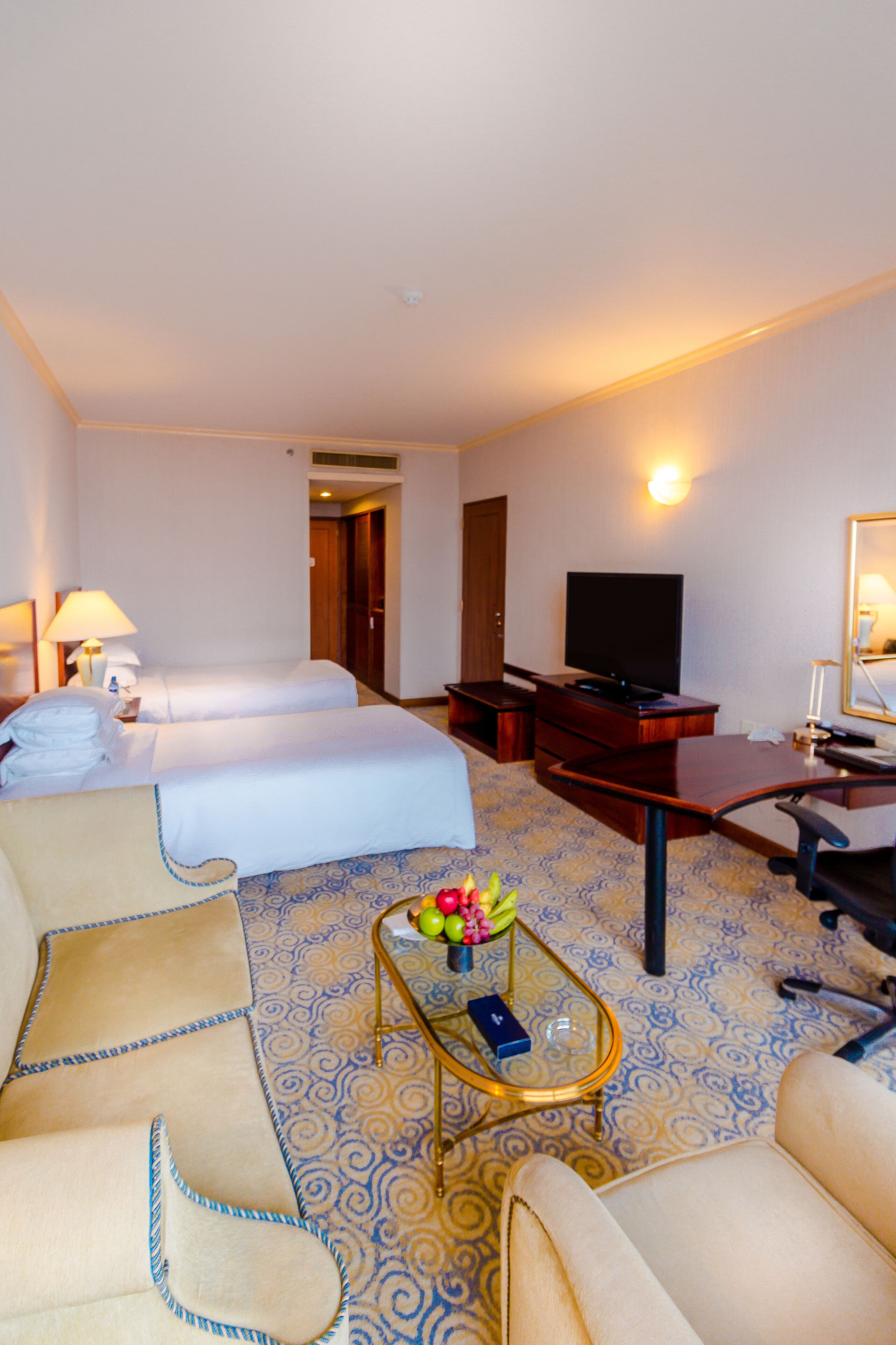 A hotel room with two beds, a desk, a television, and a small table with fruit.