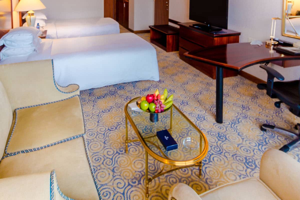 A hotel room with two beds, a desk, a television, and a small table with fruit.