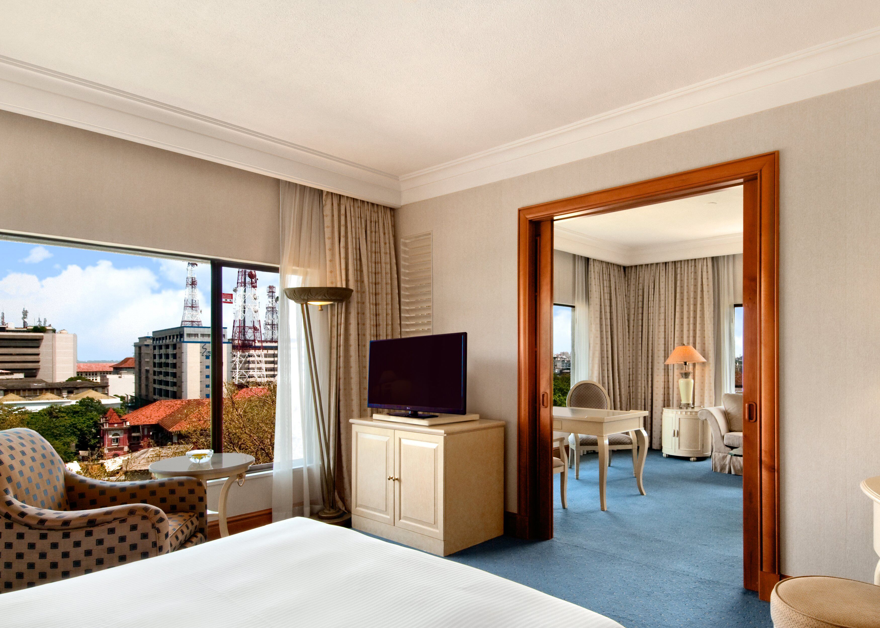 Habitación de hotel con cama, televisor, escritorio y una silla. Se observa una vista de la ciudad a través de la ventana.