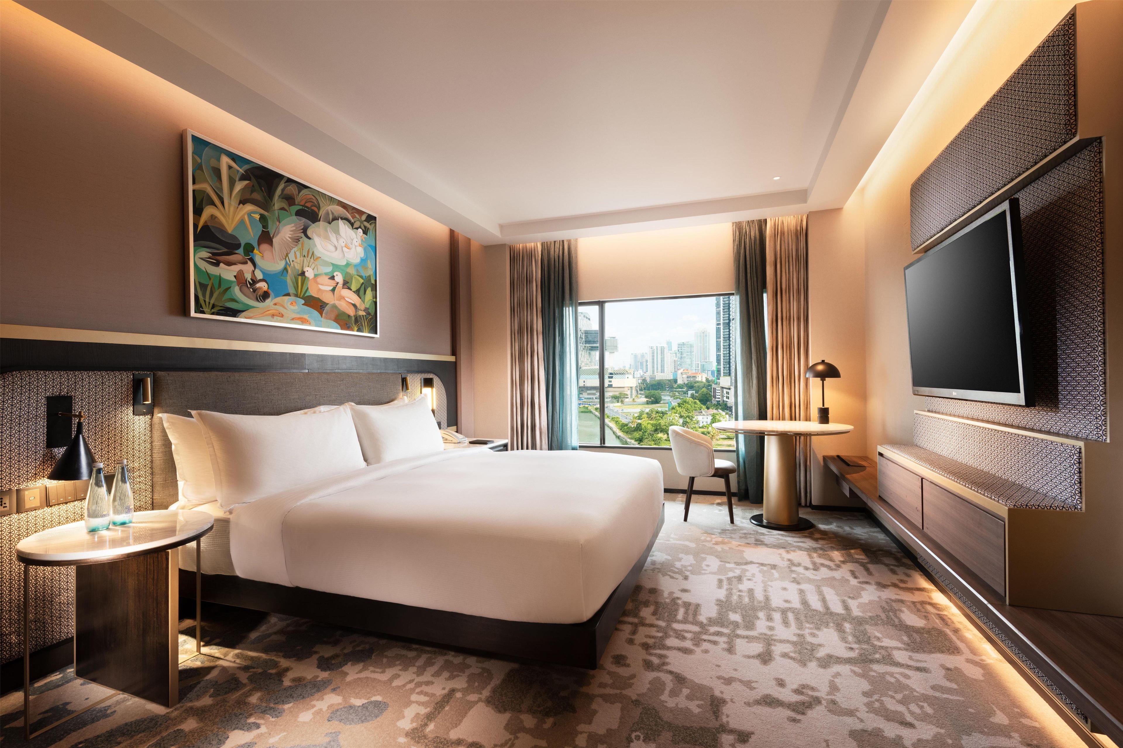 A hotel room with a large bed, a desk, a chair, a TV, and a painting on the wall.