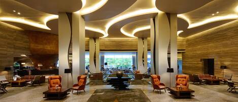 A spacious lobby with modern curved ceiling lights, a central seating area with sofas and armchairs, and a view of the outdoors through large windows.