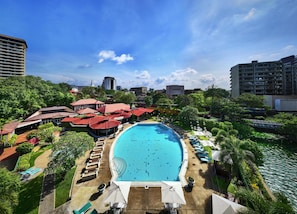 A large swimming pool surrounded by lounge chairs and umbrellas, with a view of nearby buildings and greenery.