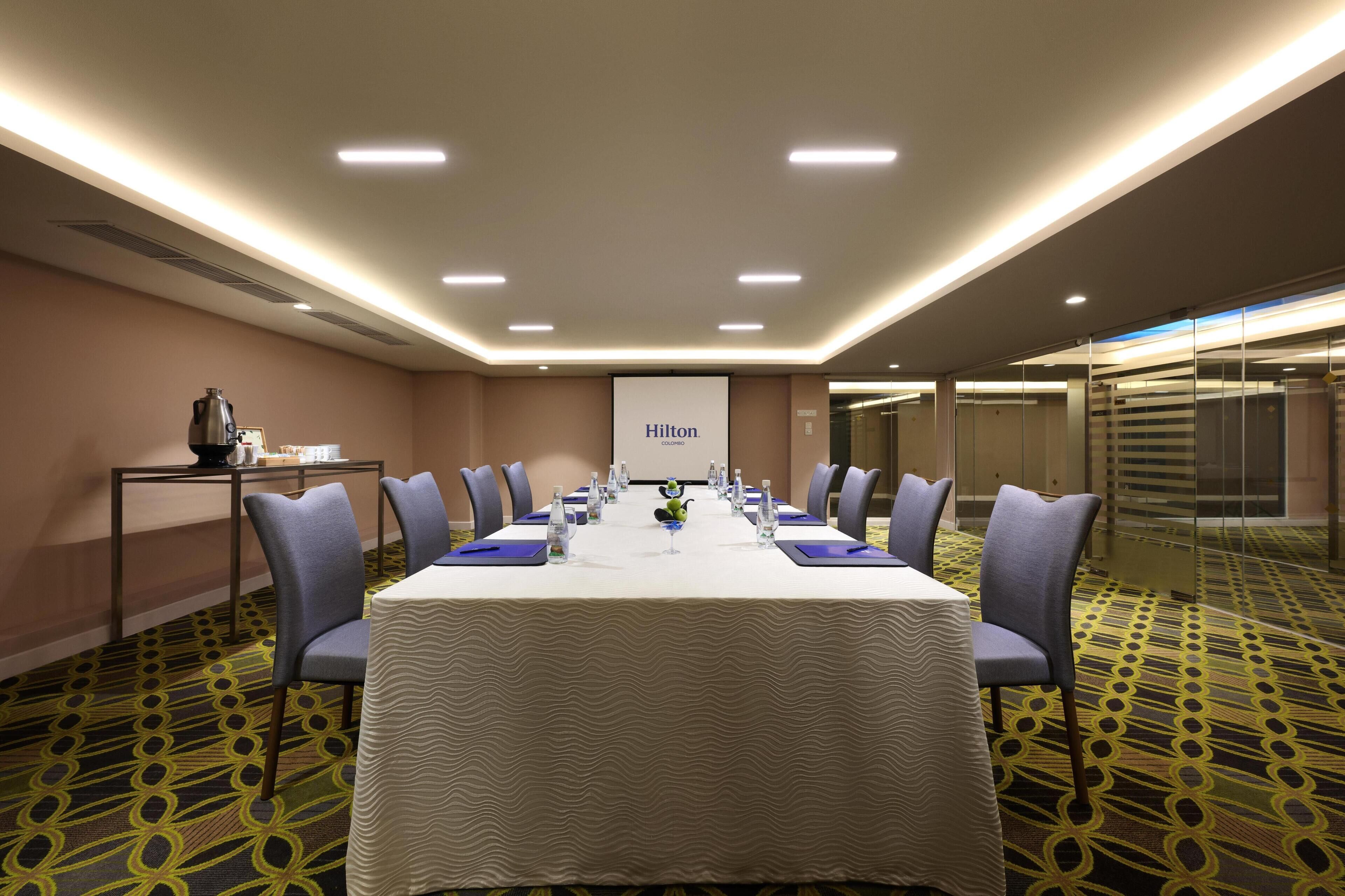 A conference room with a long table, chairs, a projector screen, and a coffee station.