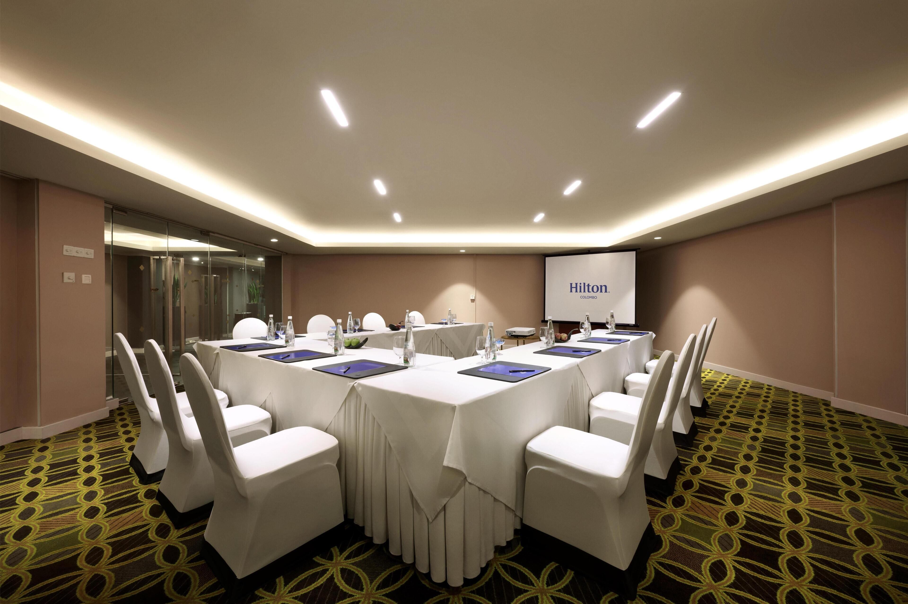 A conference room with a long table, white chairs, and a projector screen displaying "Hilton".