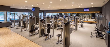 Un gimnasio moderno con diversas máquinas de ejercicio, suelo de madera y un amplio ventanal que proporciona luz natural.