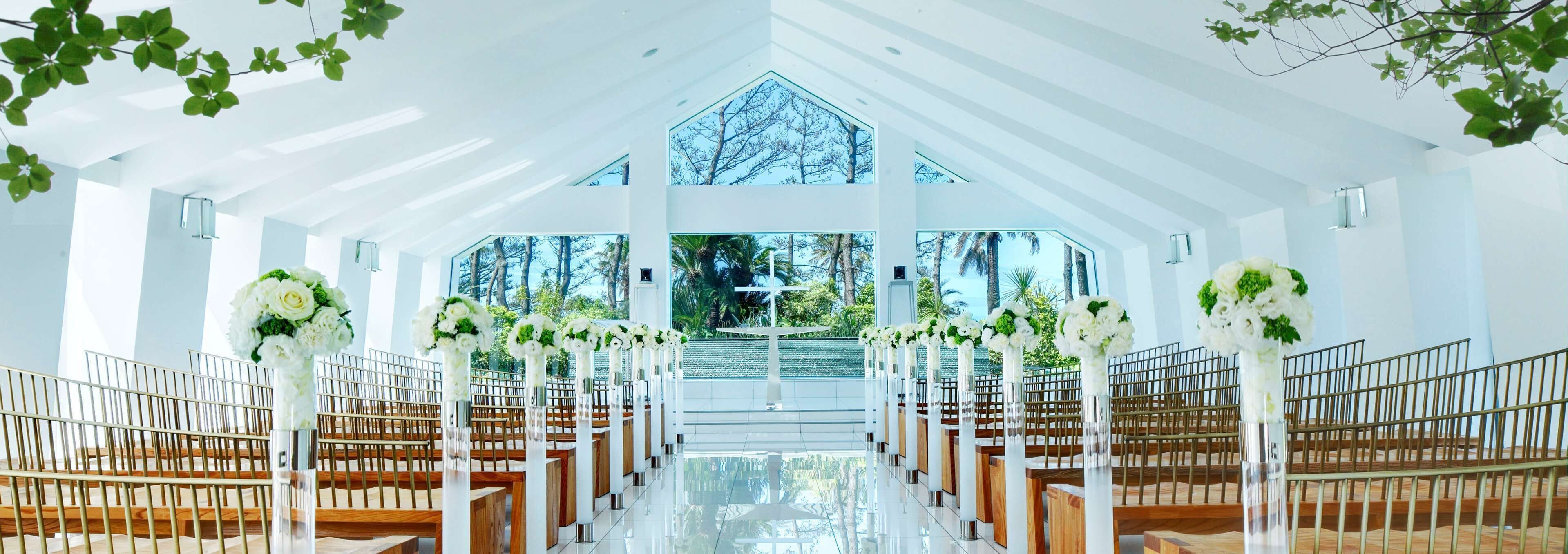 A church interior with pews arranged in rows, white floral arrangements, and a large arched window at the front.
