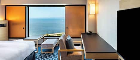 A hotel room with a bed, a desk, a chair, a TV, and a large window with a view of the ocean.