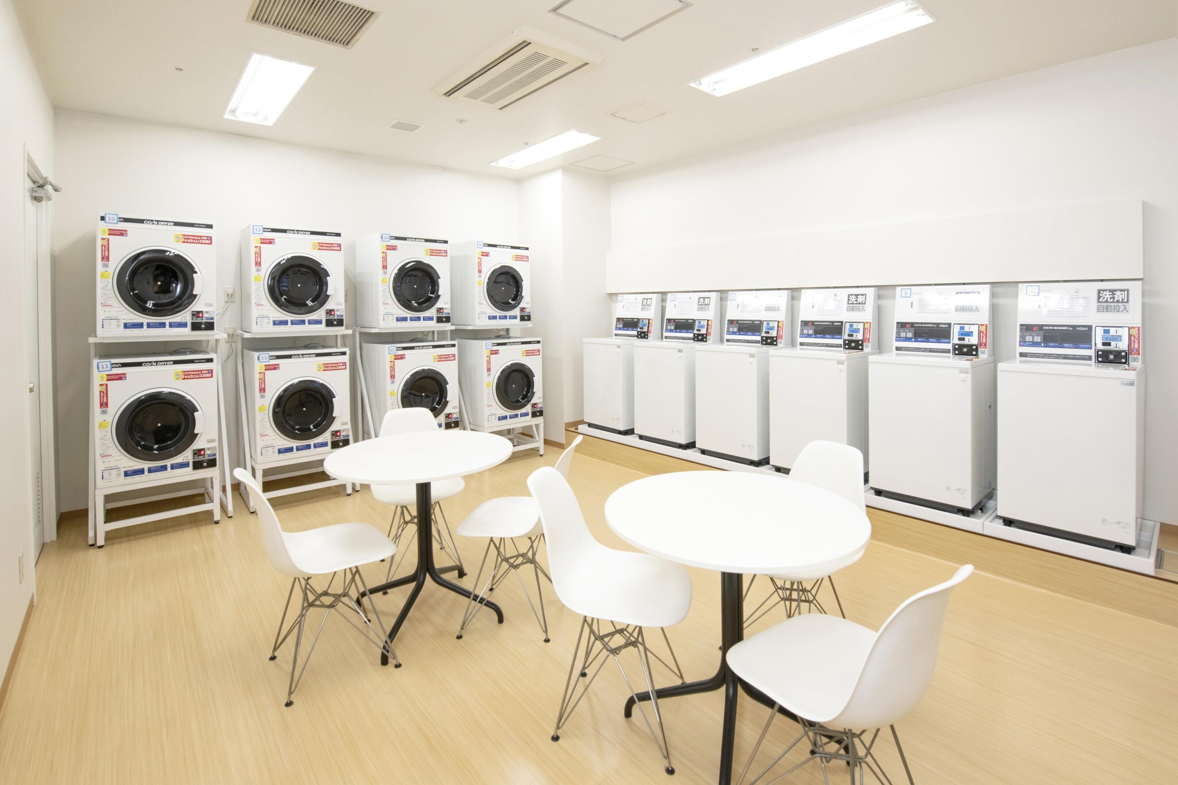 A laundry facility with multiple washing machines and dryers, white chairs, and tables.