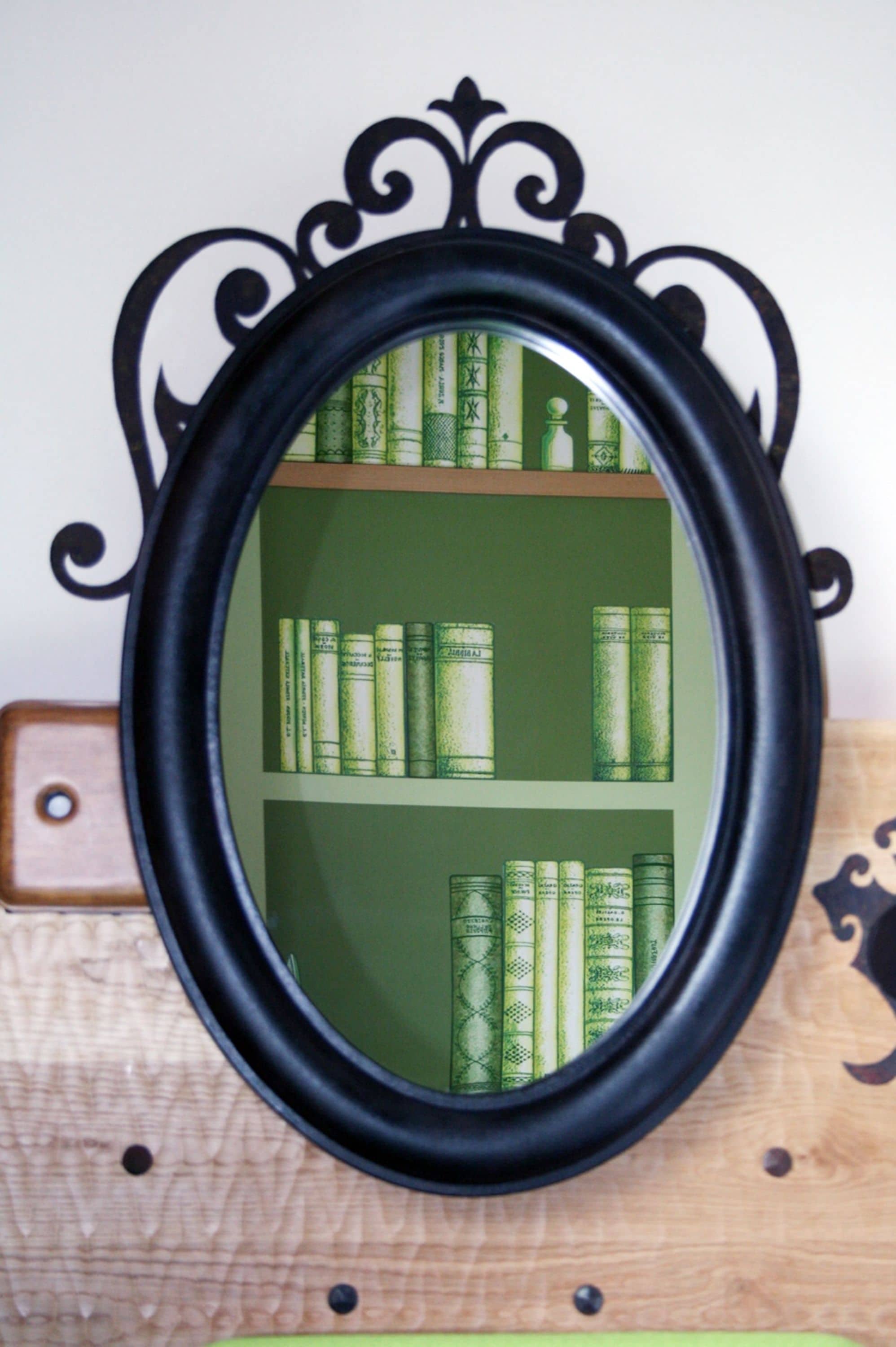 Un espejo con un marco negro ornamentado que refleja una estantería con libros verdes.