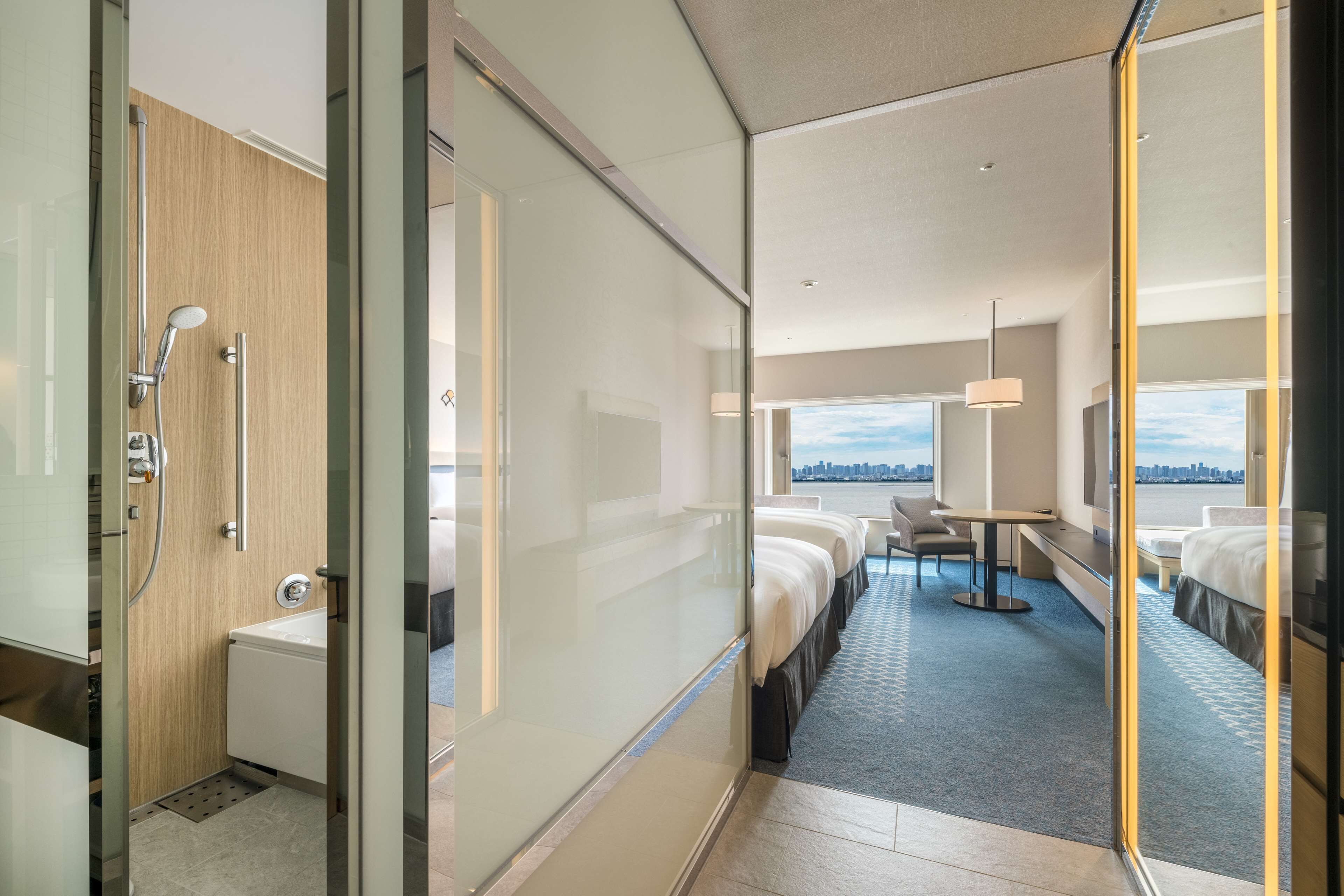 A modern hotel room with a glass partition, a shower, a bed, a desk, and a view of the city.