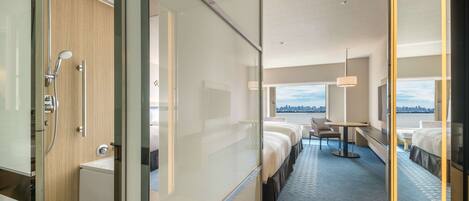 A modern hotel room with a glass partition, a shower, a bed, a desk, and a view of the city.