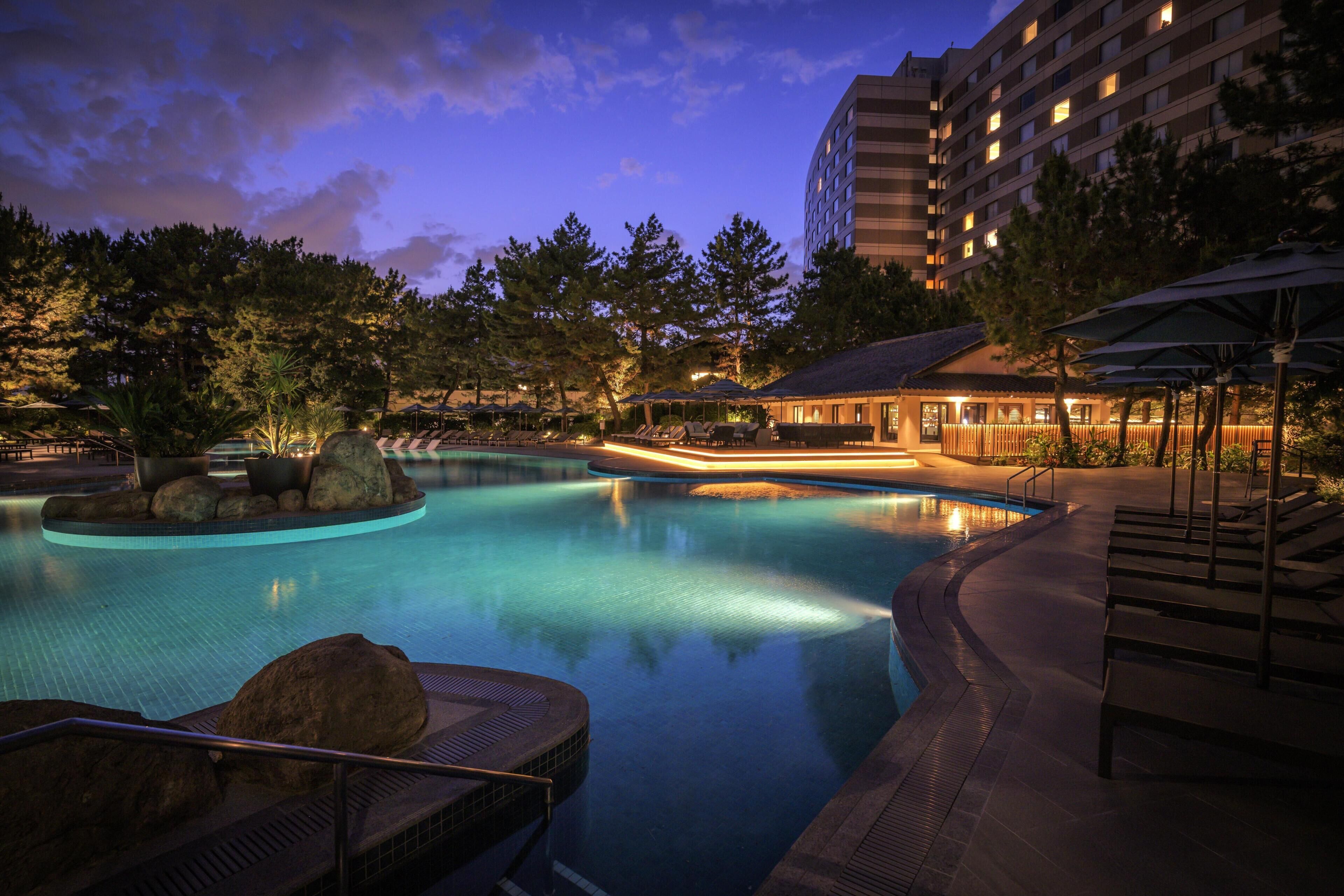 A swimming pool with lounge chairs and umbrellas, surrounded by trees and a building.