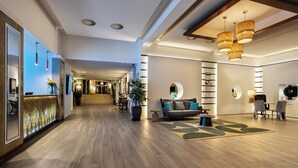 A modern hotel lobby with a wooden floor, a reception desk, a sofa with cushions, a coffee table, and a bookshelf.