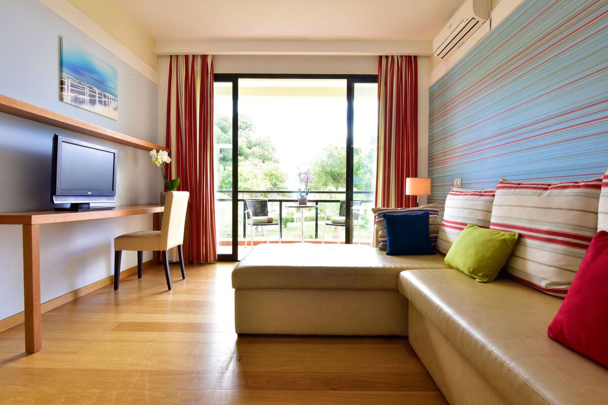 A modern hotel room with a wooden desk, a flat-screen TV, a sofa with striped cushions, and a balcony with a view of greenery.