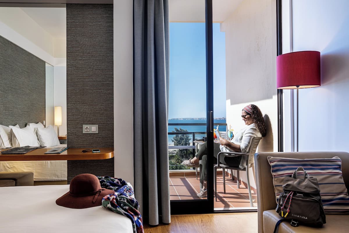 A hotel room with a bed, a desk with a lamp, a chair, a balcony with a view, and a window with curtains.