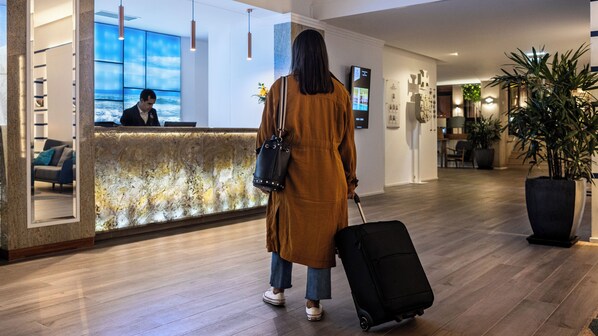 Zona de recepción de un hotel con un mostrador de mármol, una recepcionista y un huésped con una maleta.