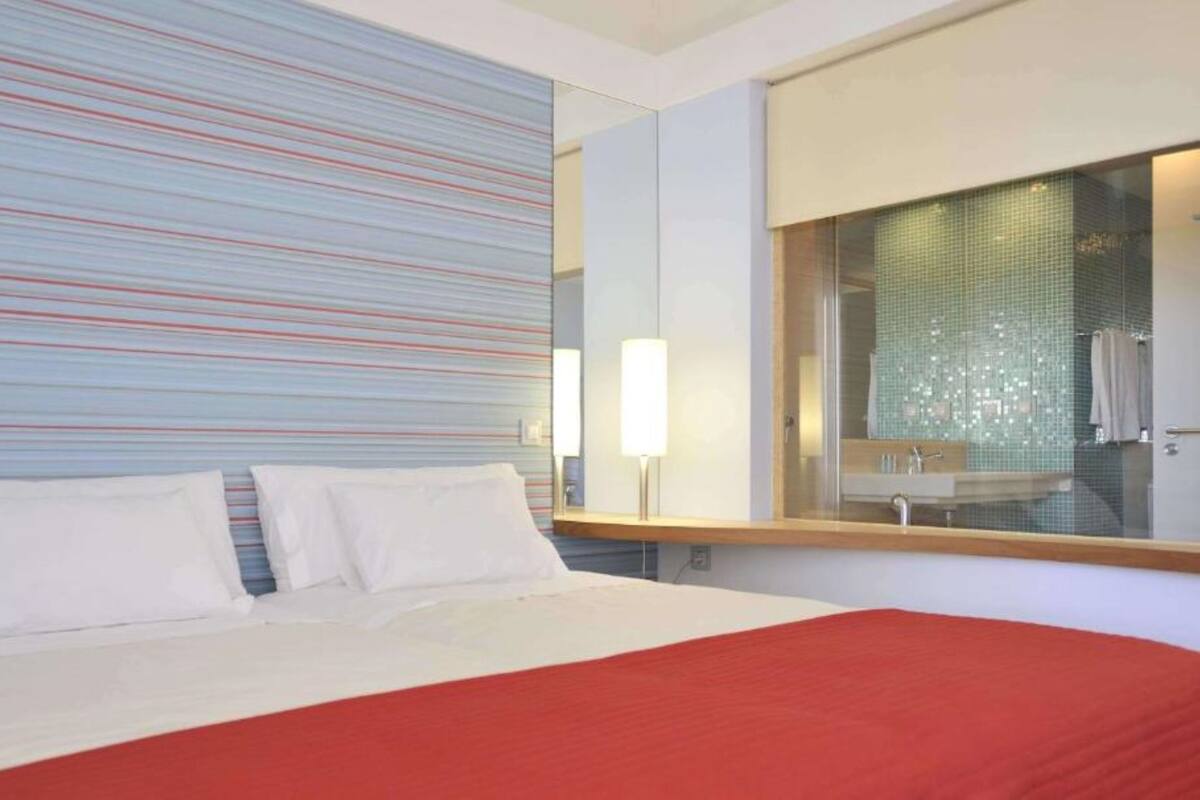 A hotel room with a large bed, a red accent wall, and a bathroom with a glass shower enclosure.