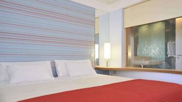 A hotel room with a large bed, a red accent wall, and a bathroom with a glass shower enclosure.