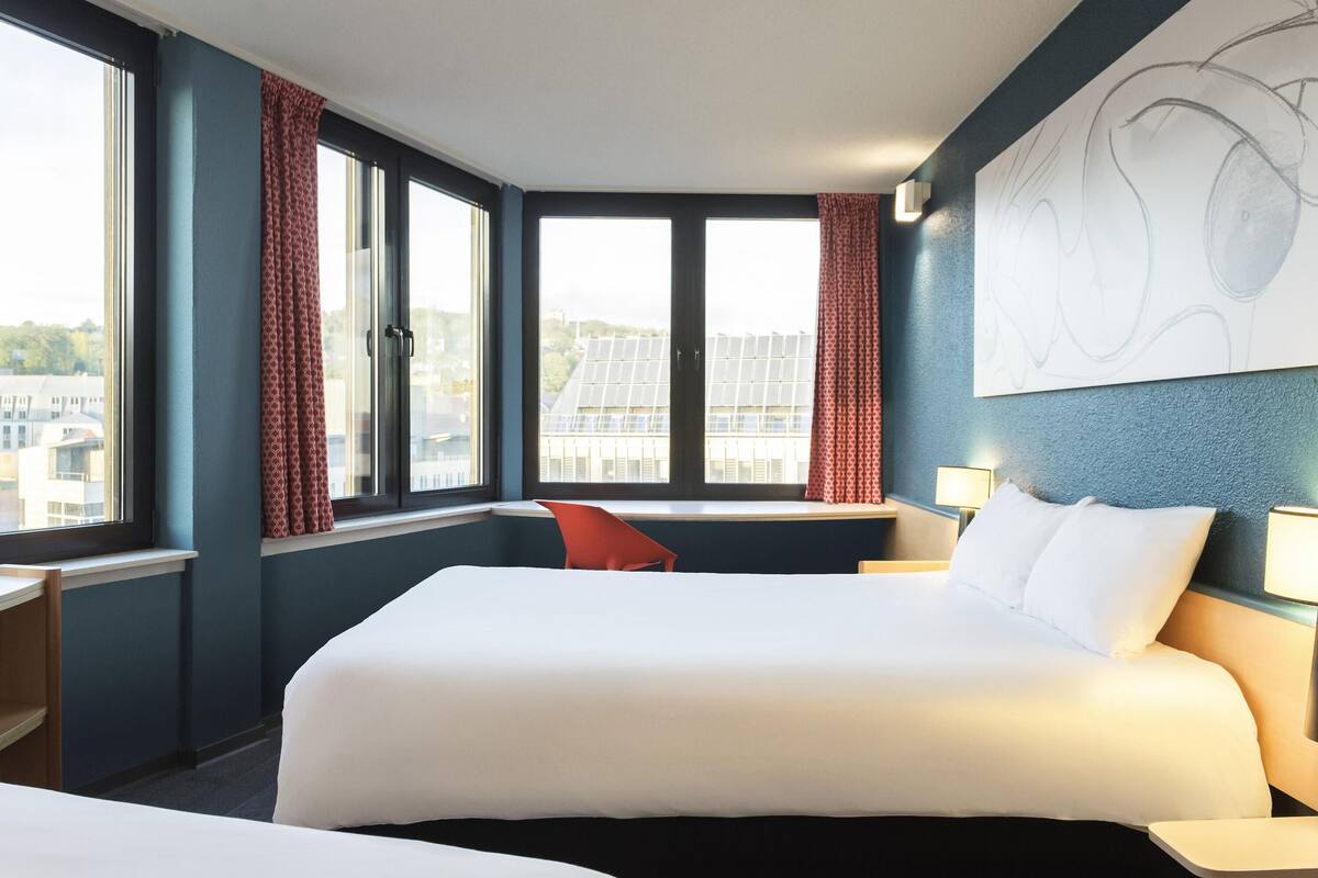 A hotel room with two beds, a desk, a chair, and large windows with curtains.