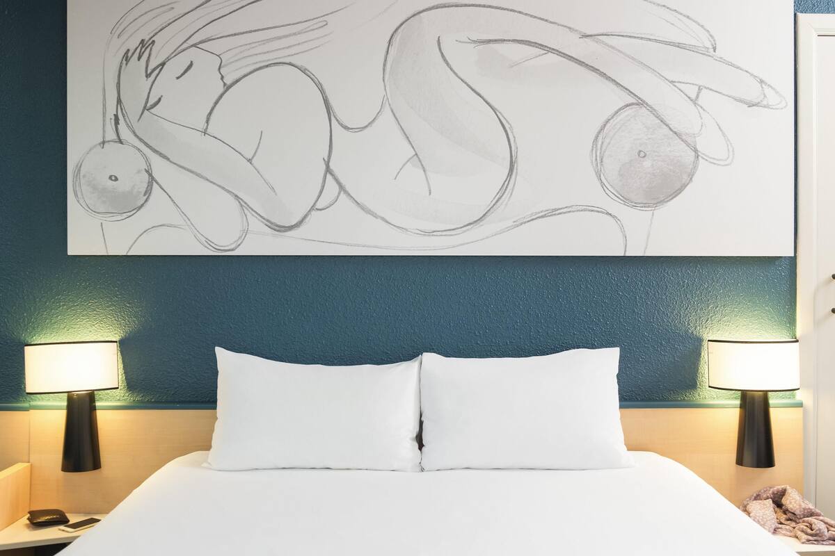 A hotel room with a bed, two bedside lamps, a wall art piece, and a door.