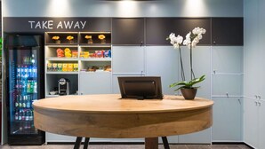 A reception area with a round wooden table, a laptop, a potted plant, and a 'TAKE AWAY' sign above a snack shelf. - ibis Liège Centre Opéra (Liege)