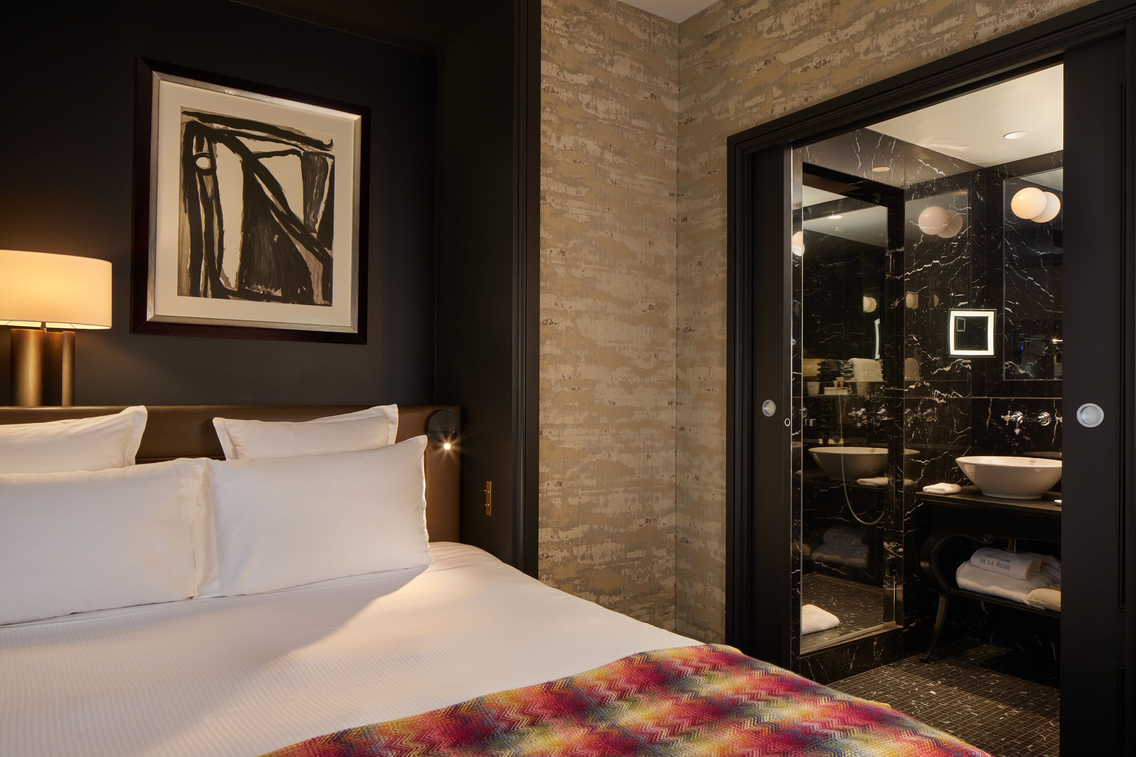 Une chambre d’hôtel comprenant un lit, un grand miroir, une salle de bain avec un lavabo et un miroir, et une œuvre d’art encadrée accrochée au mur.