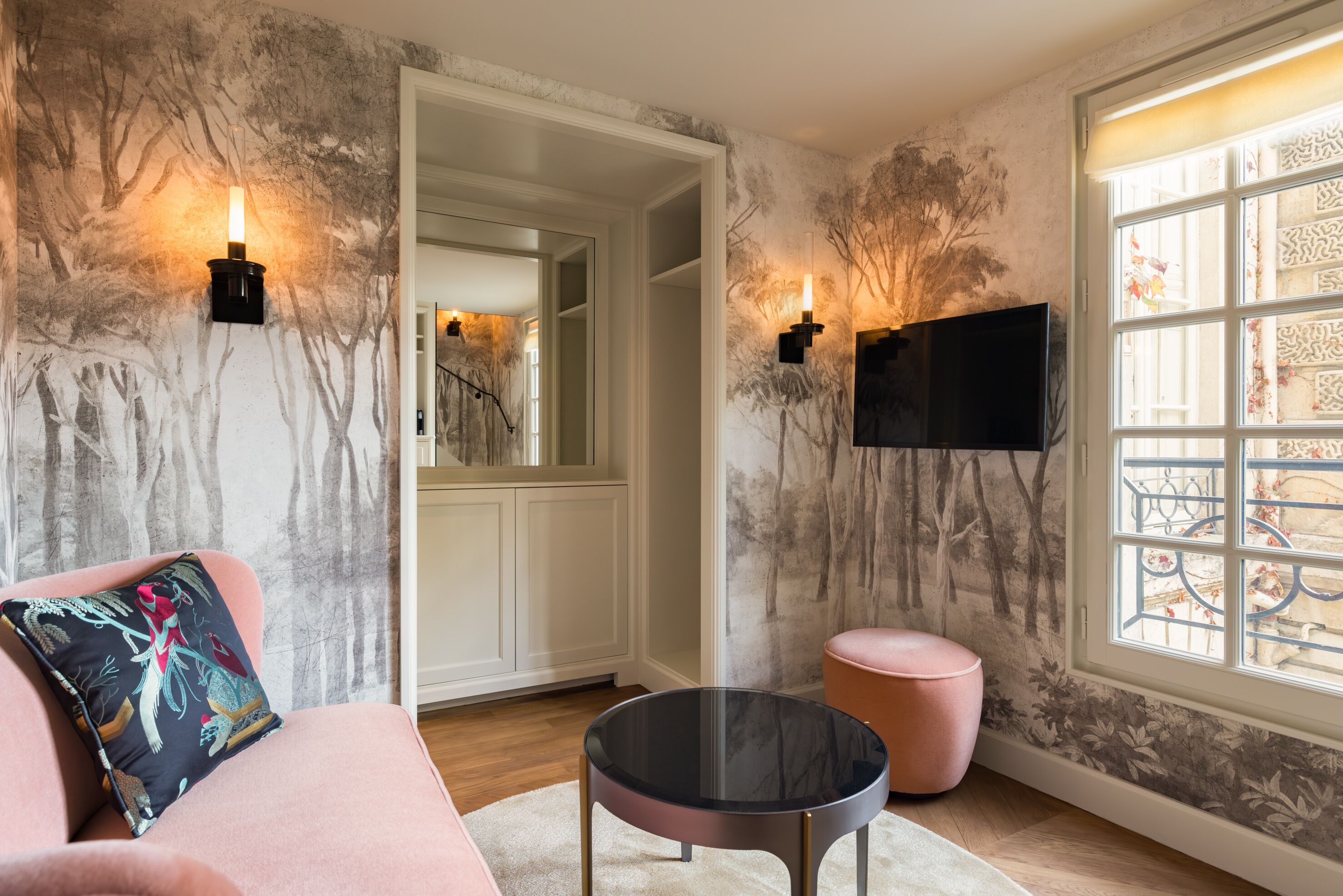 A room with a pink sofa, a round black table, a pink ottoman, and a TV mounted on the wall.