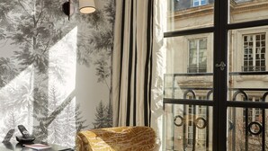 A room with a patterned armchair, a desk with a telephone, and a view of a balcony with metal railings. - Pavillon de la Reine & Spa, Place des Vosges (Paris)