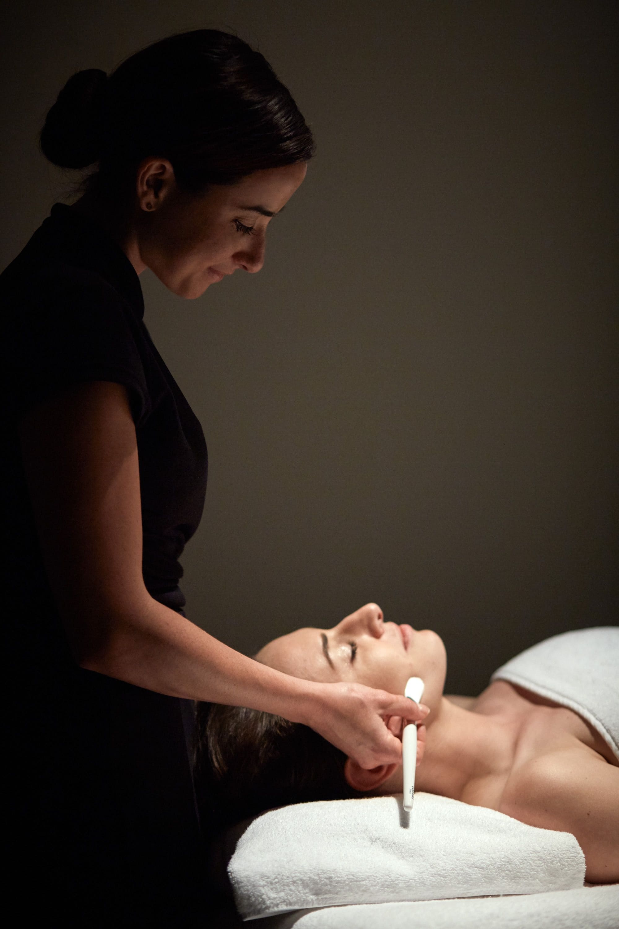 A person lying on a bed with a white towel, receiving a facial treatment from another person.