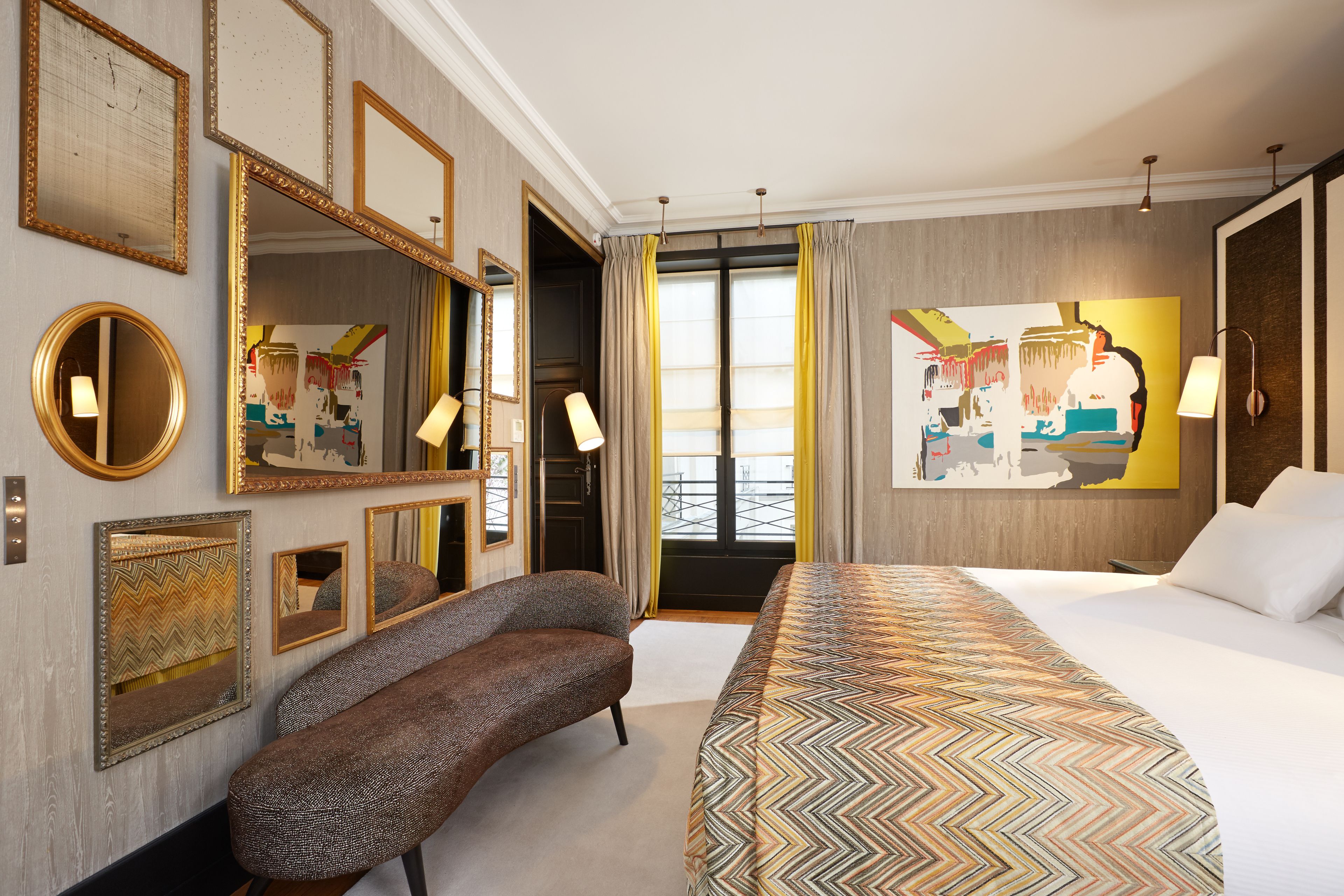 Une chambre d’hôtel avec un grand lit, un banc, plusieurs miroirs et une grande fenêtre avec des rideaux.