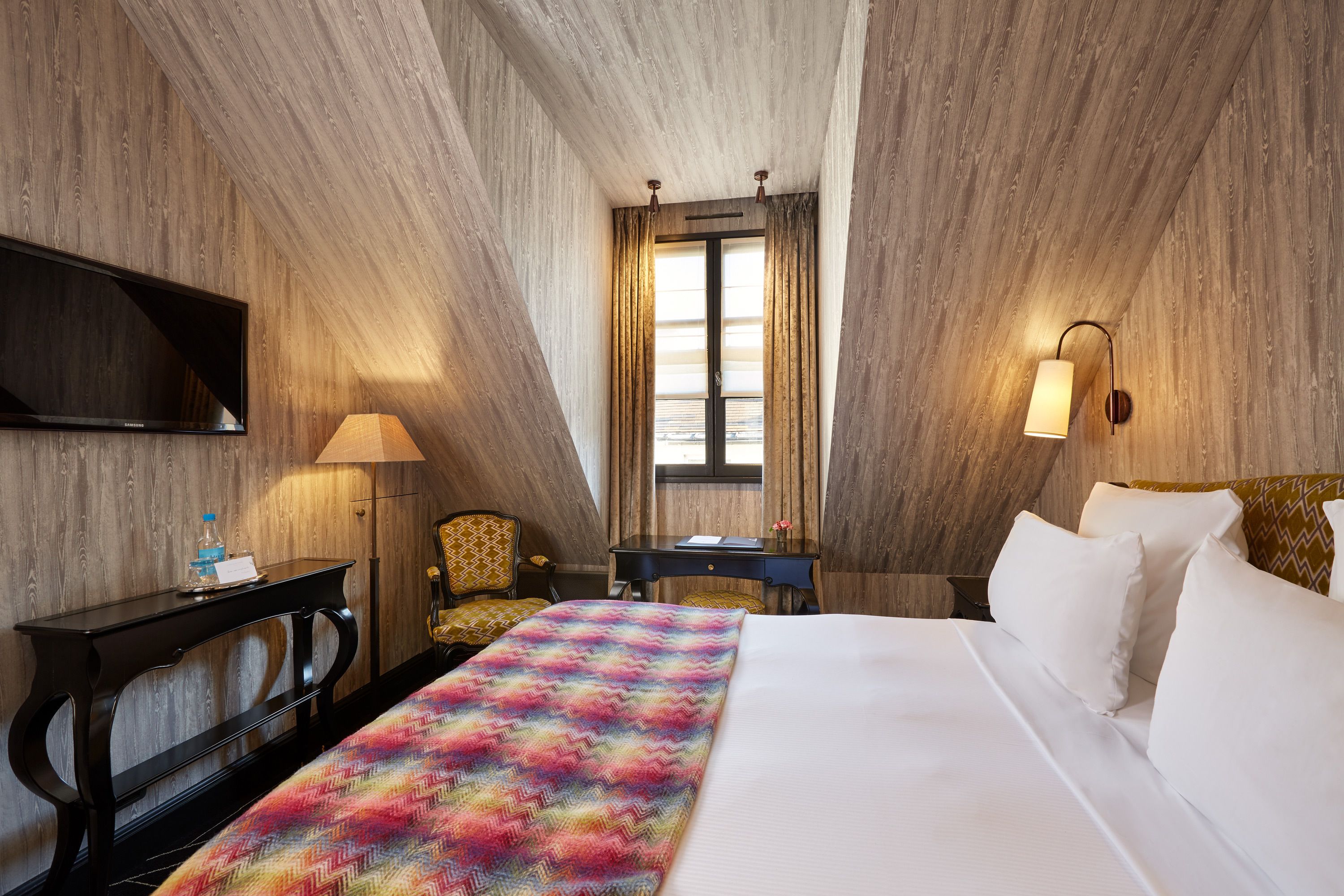 Una camera d'albergo con un letto, una scrivania, una sedia, una TV e una finestra con tende.